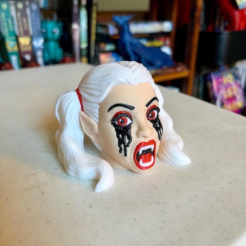 May include: A sculpted head figurine featuring white hair styled in two pigtails with red hair ties. The face has pointed ears, red eyes, black paint dripping from the eyes, and an open mouth revealing white fangs against bright red lips.