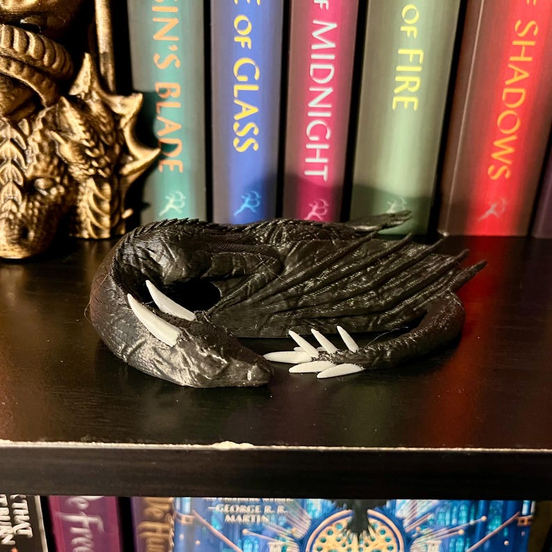 May include: A black dragon figurine with white claws, curled up and sleeping on a black shelf. The dragon is in front of a bookshelf with several books with titles like "A Song of Ice and Fire" and "A Game of Thrones".