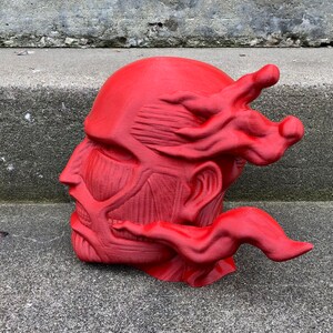 May include: A red 3D printed sculpture of a human head with a detailed muscular structure and a flowing, flame-like design on the side.