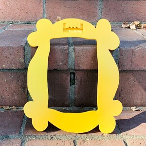 May include: A yellow plastic frame shaped like the iconic "Friends" apartment door frame. The frame has a cutout at the top for hanging.