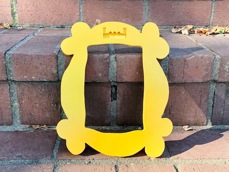 May include: A yellow plastic frame shaped like the iconic "Friends" apartment door frame. The frame has a cutout at the top for hanging.