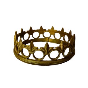 May include: A gold-colored crown with a circular design. The crown features alternating fleur-de-lis and circular cutouts around its circumference. The crown is made of metal and sits on a white background.