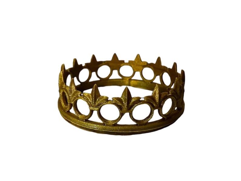 May include: A gold-colored crown with a circular design. The crown features alternating fleur-de-lis and circular cutouts around its circumference. The crown is made of metal and sits on a white background.