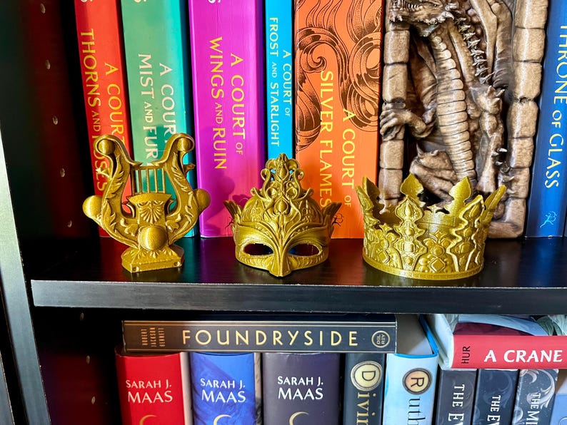 May include: Gold-colored decorative items on a black shelf, including a lyre, a masquerade mask, and a crown. The items are displayed in front of a collection of colorful books. The books have titles such as "A Court of Thorns and Roses" and "Foundryside".