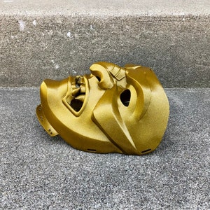 May include: A gold-coloured, detailed, plastic mask with a fierce expression. The mask is lying on a grey concrete surface.