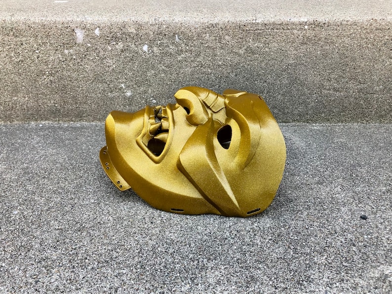 May include: A gold-coloured, detailed, plastic mask with a fierce expression. The mask is lying on a grey concrete surface.