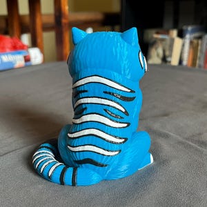 Puede incluir: Figura de gato azul con rayas blancas y negras. El gato est&aacute; sentado con la cola enroscada y las orejas erguidas. La figura est&aacute; hecha de un material liso con un acabado brillante. Los ojos del gato son blancos con pupilas negras.