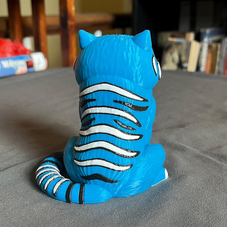 Puede incluir: Figura de gato azul con rayas blancas y negras. El gato est&aacute; sentado con la cola enroscada y las orejas erguidas. La figura est&aacute; hecha de un material liso con un acabado brillante. Los ojos del gato son blancos con pupilas negras.