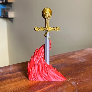 May include: A decorative sword figurine with a gold hilt and a silver blade, set against a vibrant red flame base. The hilt features an ornate design, and the flame base adds a dramatic flair. This fantasy-themed item is suitable for display.