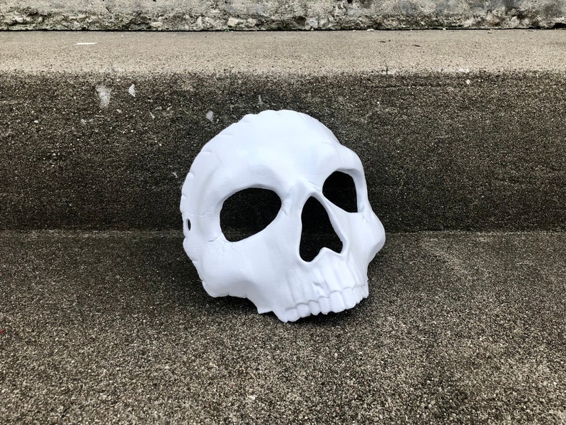 Peut inclure: Un masque de cr&acirc;ne blanc avec de grandes orbites oculaires et une cavit&eacute; nasale pro&eacute;minente. Le masque est fait d'un mat&eacute;riau lisse et repose sur une surface grise textur&eacute;e. Le masque est con&ccedil;u pour &ecirc;tre port&eacute;.
