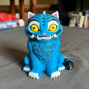 Puede incluir: Una figura azul de una criatura felina con grandes ojos amarillos, colmillos blancos y marcas negras. La figura tiene un patr&oacute;n de manchas en el pecho y una cola a rayas. Las orejas y la boca son rosas.