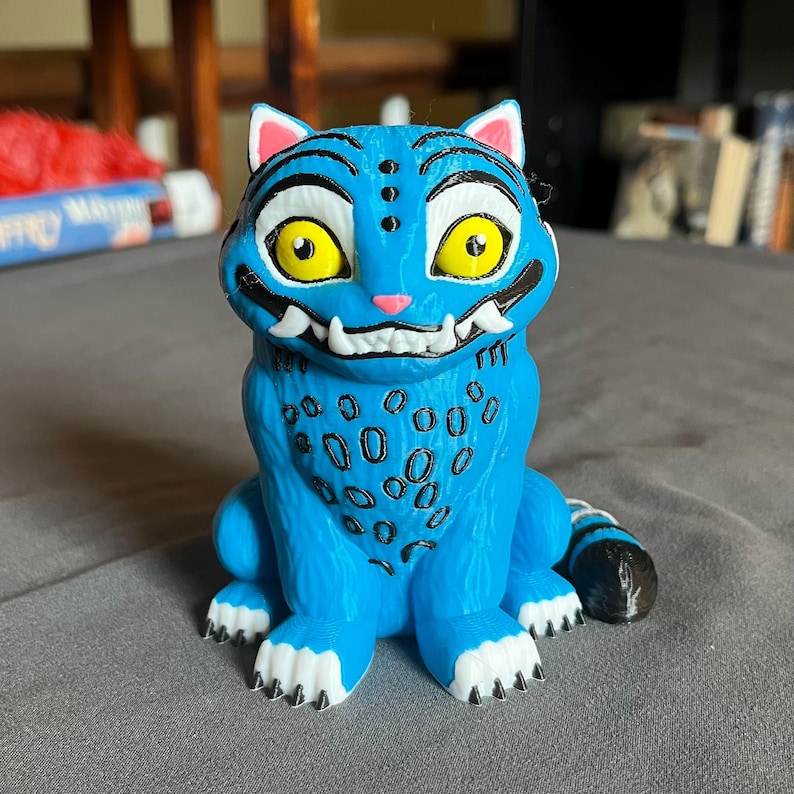 Puede incluir: Una figura azul de una criatura felina con grandes ojos amarillos, colmillos blancos y marcas negras. La figura tiene un patr&oacute;n de manchas en el pecho y una cola a rayas. Las orejas y la boca son rosas.