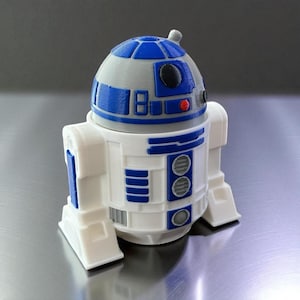 Może przedstawiać: Figurka R2-D2 wydrukowana w 3D. Robot-zabawka ma biały korpus z niebieskimi i szarymi detalami, w tym niebieską kopułkową głowę. Posiada prostokątne i okrągłe elementy konstrukcyjne z małym czerwonym akcentem.