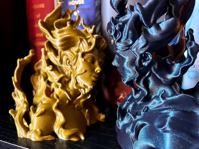 May include: Two detailed busts, one gold and one dark blue, face each other. The busts feature flowing, flame-like hair and classical facial features. The gold bust is on the left, and the dark blue bust is on the right.