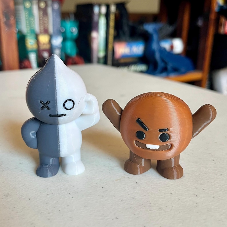 May include: Two 3D-printed figurines. One is gray and white, with an eye and an X on its face. The other is brown with a round body, arms raised, and a stern expression. The figurines are on a light-colored surface.
