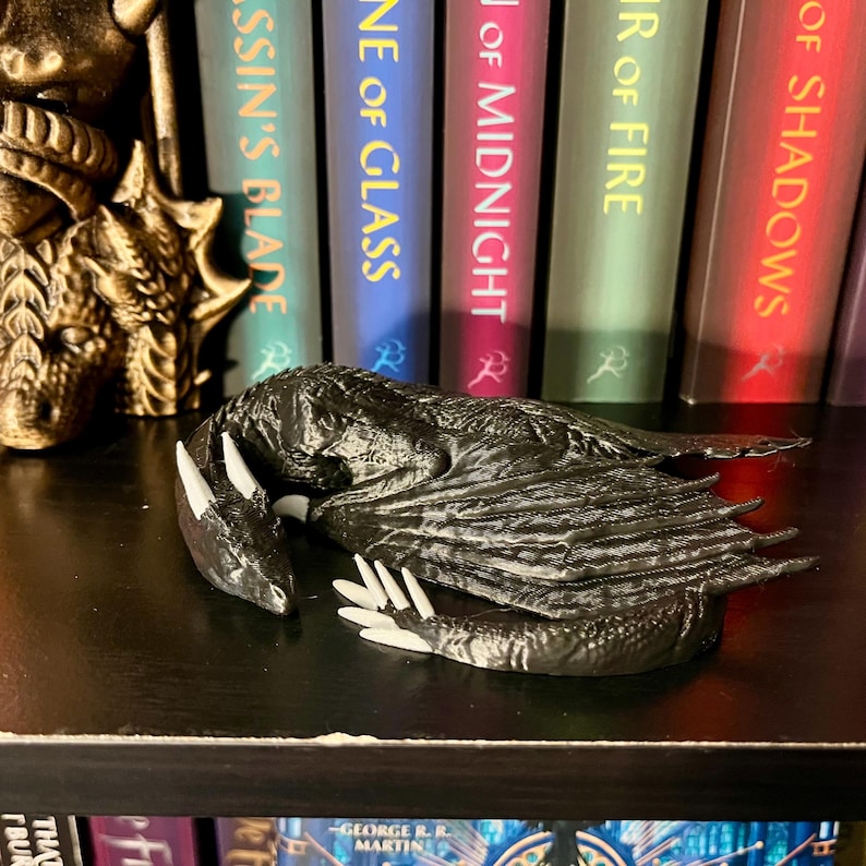 May include: A black dragon figurine curled up in a sleeping position. The dragon has white claws and a white underbelly. The figurine is on a shelf with a row of books behind it.