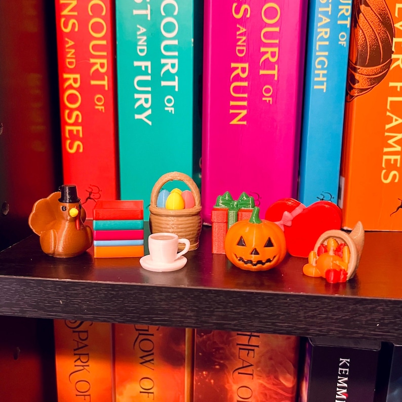 Op de afbeelding: Een plank toont miniatuurbeeldjes met een vakantiethema. Een bruine kalkoen met hoge hoed, een stapel kleurrijke blokken, een mand met paaseieren, een kopje koffie, een pompoen en een hartvormige decoratie zijn zichtbaar.
