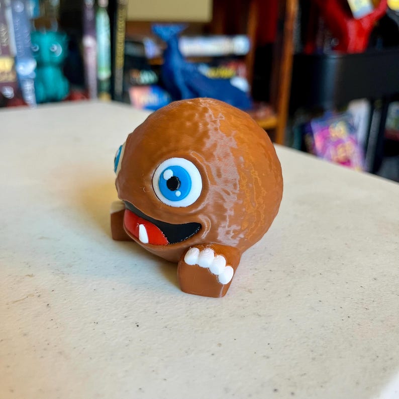 Puede incluir: Una figura de criatura esf&eacute;rica de color marr&oacute;n impresa en 3D con un cuerpo liso y redondeado. Presenta un ojo grande de color azul brillante con una pupila negra, una boca ancha negra con un solo diente blanco y pies peque&ntilde;os de color marr&oacute;n con dedos blancos.
