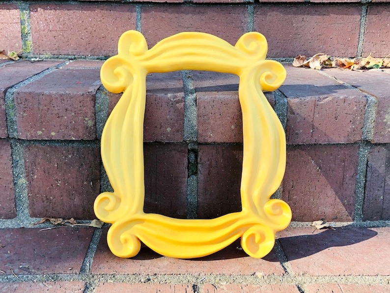 May include: A yellow plastic frame with a unique, wavy design. The frame is shaped like the iconic frame from the TV show "Friends".