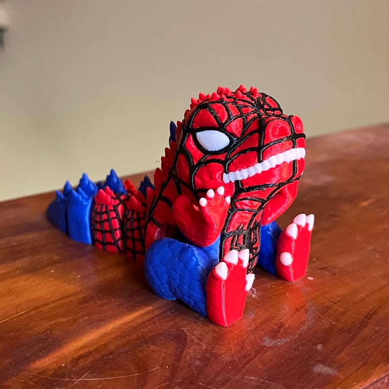 May include: A red, blue, and black 3D-printed figurine of a dinosaur-like creature. The figure has a red body with black web-like patterns, blue accents, and white teeth and eyes. The figurine is sitting on a wooden surface.