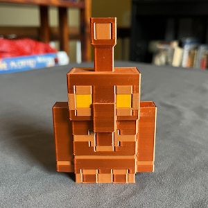 Könnte beinhalten: Eine braune, pixelige Statue einer Kreatur mit rechteckigem Körper, Armen und Kopf. Die Augen sind gelb und die Statue hat einen hohen, rechteckigen Vorsprung auf dem Kopf. Die Statue besteht aus einem 3D-gedruckten Material.