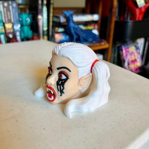May include: A small figurine of a head with white hair styled in two ponytails tied with red bands. The face features pale skin, red eyes, black makeup dripping from the eyes, and an open mouth showing white fangs. The ears are pointed.