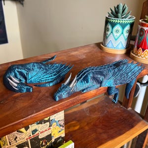 Puede incluir: Dos dragones impresos en 3D de color azul turquesa, uno enroscado y el otro acostado boca abajo con la cabeza girada hacia un lado, se exhiben en un estante de madera.
