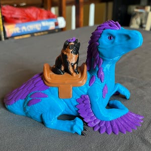 Puede incluir: Una figura de dinosaurio azul y morado con una silla de montar y una peque&ntilde;a figura de perro con una corona. El dinosaurio tiene un dise&ntilde;o de plumas y est&aacute; sentado. El perro es negro y marr&oacute;n.