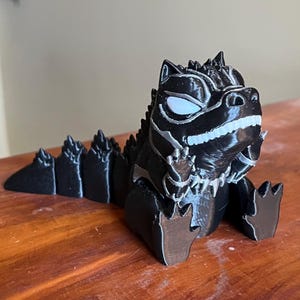 May include: A black and silver 3D-printed figurine of a stylized dragon or dinosaur. The figurine has a detailed design with spikes along its back and a smiling expression. The eyes are white, and the figurine is sitting down.