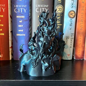 May include: A dark blue sculpture of a person's bust with stylized flames. The sculpture is on a shelf in front of books with titles including "Crescent City" and "House of Sky and Breath". The sculpture's design evokes a fiery aesthetic.