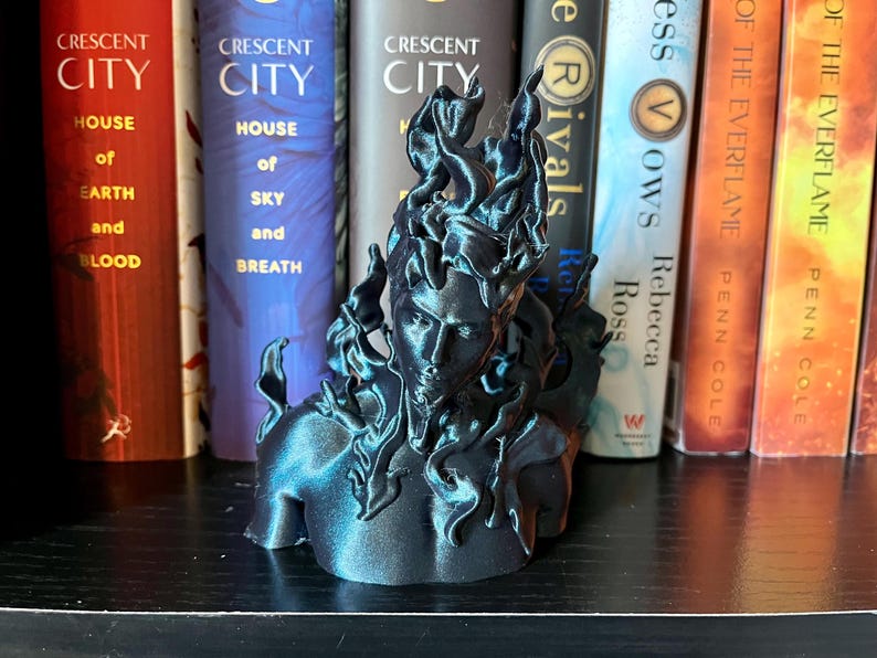 May include: A dark blue sculpture of a person's bust with stylized flames. The sculpture is on a shelf in front of books with titles including "Crescent City" and "House of Sky and Breath". The sculpture's design evokes a fiery aesthetic.