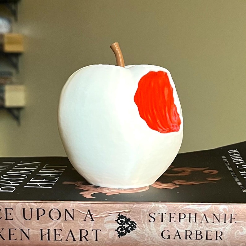 Puede incluir: Una escultura blanca con forma de manzana con un mordisco que revela un interior rojo brillante. La escultura tiene un tallo marr&oacute;n y est&aacute; colocada sobre un libro titulado "Once Upon a Broken Heart".