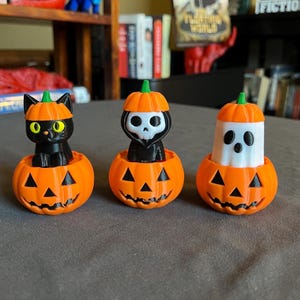 May include: Three Halloween-themed figurines: a black cat, a grim reaper, and a ghost, each inside an orange pumpkin. The pumpkins have carved jack-o'-lantern faces. The cat has yellow eyes, and the ghost has black eyes and mouth.