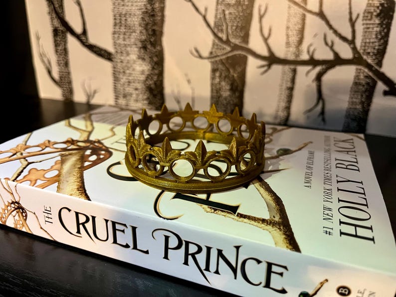 May include: A gold-colored crown with fleur-de-lis details sits atop a book titled "The Cruel Prince" by Holly Black. The book has a white cover with illustrations. The background features a black and white patterned design.