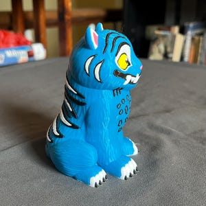 Puede incluir: Una figura de tigre azul con rayas negras y detalles blancos. El tigre tiene un ojo amarillo, colmillos blancos y garras negras. La figura est&aacute; sobre una superficie gris.