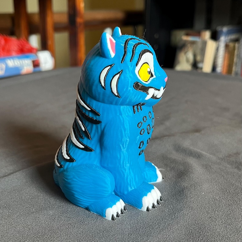 Puede incluir: Una figura de tigre azul con rayas negras y detalles blancos. El tigre tiene un ojo amarillo, colmillos blancos y garras negras. La figura est&aacute; sobre una superficie gris.