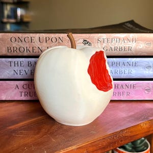 Puede incluir: Una escultura de cerámica blanca con forma de manzana a la que le falta un mordisco, revelando un interior rojo brillante. La manzana tiene un tallo marrón y está sobre una superficie de madera. Libros con títulos como "Once Upon Broken" están en el fondo.