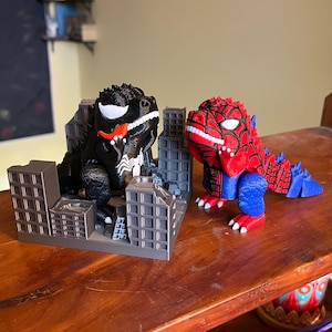 May include: Two 3D-printed dinosaur figures, one black and one red, each with superhero-themed designs. The black figure resembles Venom, with a white emblem and red tongue. The red figure is Spider-Man themed. Both are posed in front of miniature cityscapes.