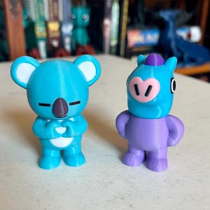 May include: Two 3D-printed figurines. One is a light blue koala with a brown nose and closed eyes, hands clasped in a heart shape. The other is a purple figure with a blue head, pink snout, and a purple hat.