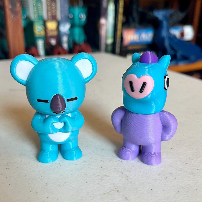 May include: Two 3D-printed figurines. One is a light blue koala with a brown nose and closed eyes, hands clasped in a heart shape. The other is a purple figure with a blue head, pink snout, and a purple hat.
