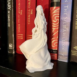 May include: White 3D printed figurine of a woman with long hair wearing a flowing gown. The figurine is standing on a shelf with several books behind it. The books are all red and have gold lettering on the spines. The titles of the books are "The Fellowship of the Ring", "The Two Towers", "The Hobbit", and "The Silmarillion".