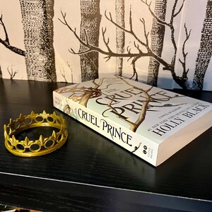 May include: A gold-colored crown and a copy of "The Cruel Prince" by Holly Black are displayed on a black surface. The book cover features a tree design. The crown is circular with decorative points.