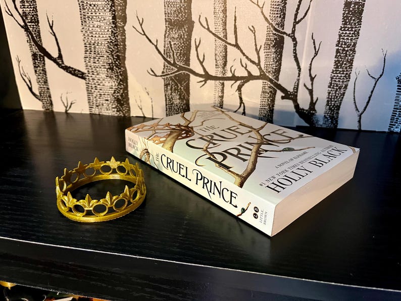 May include: A gold-colored crown and a copy of "The Cruel Prince" by Holly Black are displayed on a black surface. The book cover features a tree design. The crown is circular with decorative points.