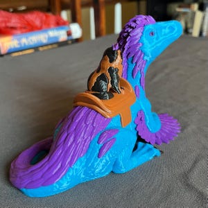 Puede incluir: Figura de dinosaurio impresa en 3D en tonos azules y morados, con una peque&ntilde;a figura de gato montada en su espalda. El gato es negro y marr&oacute;n, y la silla de montar es marr&oacute;n. El dinosaurio tiene una cola larga y est&aacute; sentado.
