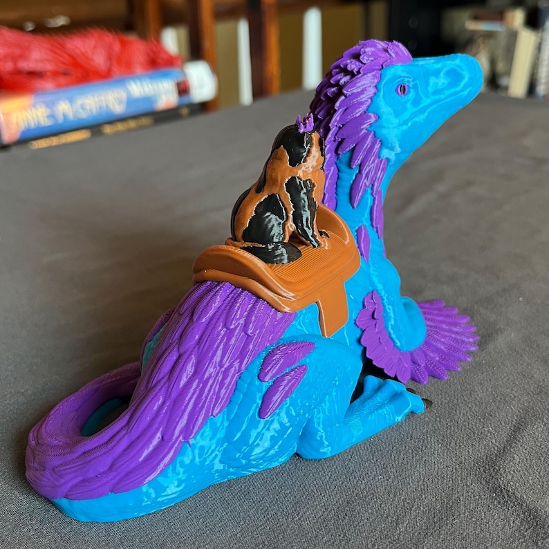 Puede incluir: Figura de dinosaurio impresa en 3D en tonos azules y morados, con una peque&ntilde;a figura de gato montada en su espalda. El gato es negro y marr&oacute;n, y la silla de montar es marr&oacute;n. El dinosaurio tiene una cola larga y est&aacute; sentado.