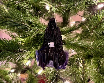 Op de afbeelding: Zwarte kerstversiering in de vorm van een figuur in een gekapte mantel die een wit theekopje vasthoudt. De versiering hangt aan een groene kerstboom.