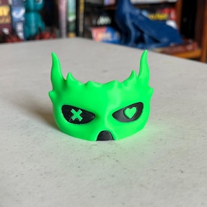K&ouml;nnte beinhalten: Eine leuchtend gr&uuml;ne Maske mit stilisiertem Gesicht. Die Maske hat schwarze Augenh&ouml;hlen, eine mit einem "X" und die andere mit einem Herzen. Die Maske hat zwei spitze H&ouml;rner und eine kleine Nase.