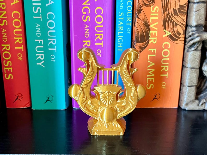 May include: A gold-colored lyre figurine, a classical musical instrument, stands in front of a row of colorful books. The lyre has an ornate design with a detailed base and strings. The books have titles like "A Court of Silver Flames".