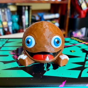 May include: A brown, spherical, 3D-printed figurine with a wide, grinning mouth, a bright red tongue, and a single white tooth. The character features large, round, light blue eyes with black pupils and small, white, three-fingered hands.