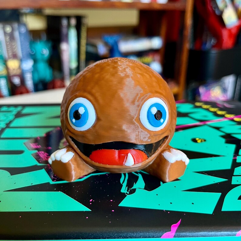 May include: A brown, spherical, 3D-printed figurine with a wide, grinning mouth, a bright red tongue, and a single white tooth. The character features large, round, light blue eyes with black pupils and small, white, three-fingered hands.